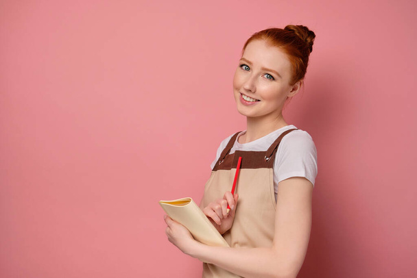 A red-haired girl in an apron with collected hair is standing on a pink background in half-turn with a notebook and smiles cute. - Fotografie, Obrázek