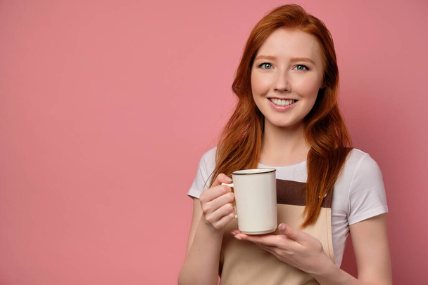 The red-haired girl in an apron stands on a pink background with a white cup in her hand and smiles broadly - Foto, Imagen