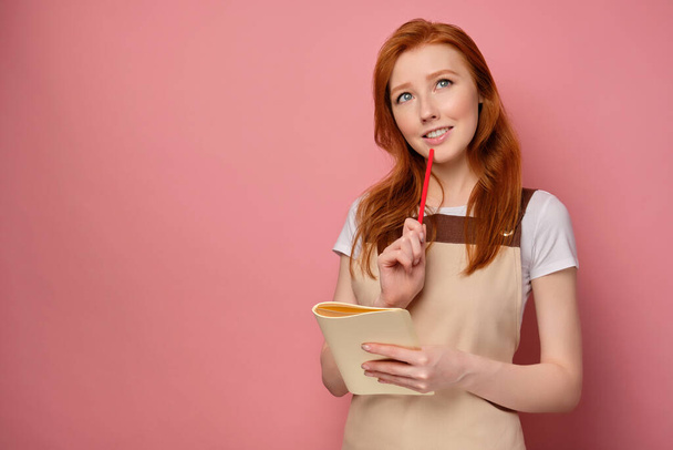 The red-haired girl in an apron stands on a pink background with a notebook and with a pencil on her lips, smiles and looks up - Fotografie, Obrázek