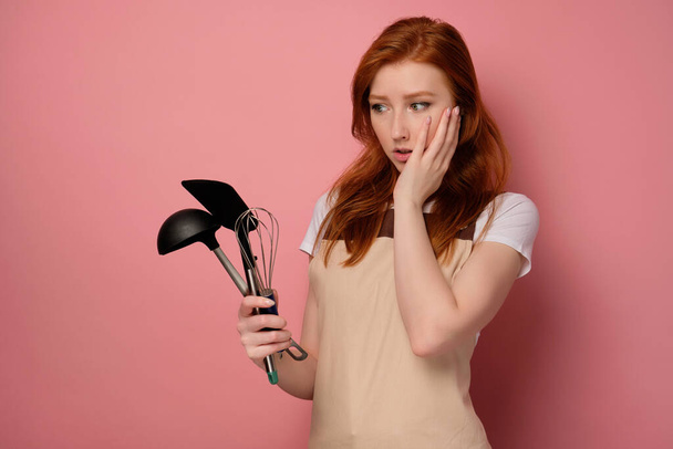 A red-haired girl in an apron stands on a pink background and looks at bewildered at the kitchen appliances in his hand - Photo, Image
