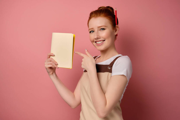 A red-haired girl in an apron with collected hair stands in a half-coat on a pink background and points a finger to the notebook - Photo, image