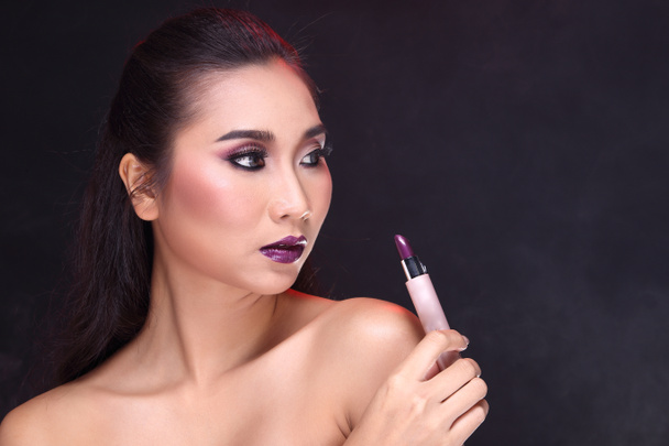 Beauty fashion portrait of Asian black hair woman with purple lipstick and hand holding lipstick near her mouth and look at stick, black background with hair lightt, copy space for text - Photo, Image