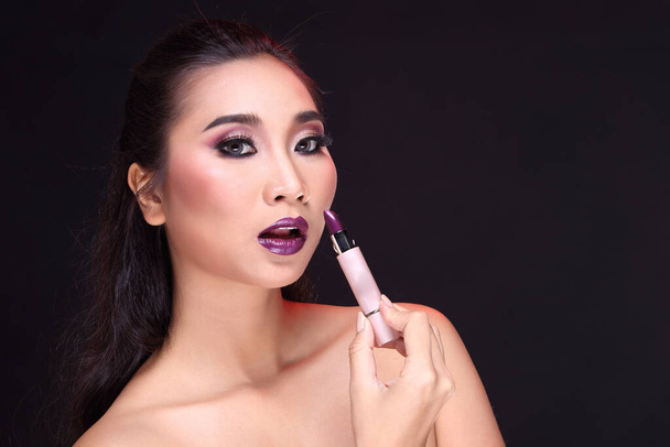 Beauty fashion portrait of Asian black hair woman with glossy purple lipstick and hand holding lipstick near her mouth, black background with hair lightt, copy space for text - Photo, Image