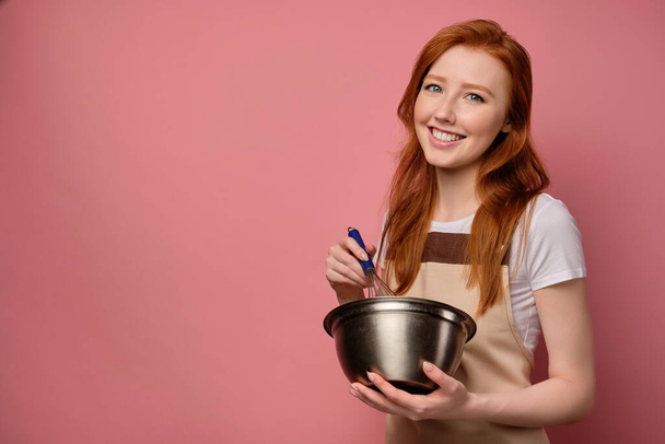 A red-haired girl in an apron stands on a pink background and whips with a whisk in a bowl and smiles broadly into the frame - Valokuva, kuva