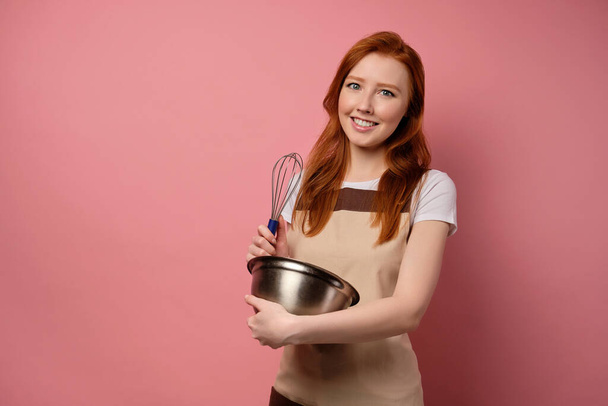The red-haired girl in an apron stands on a pink background with a whisk and a bowl - Фото, изображение