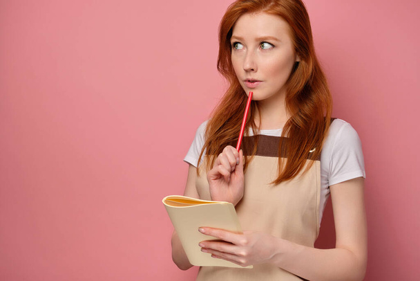 The red-haired girl in an apron is standing on a pink background with a notebook and looking at the side with a pencil to her lips - Фото, зображення