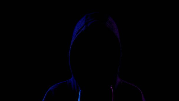 Молодая женщина стоит в капюшоне, скрывая свое лицо. Нападавший стоит в темноте. Синий и красный свет падает на человека на черном фоне
. - Фото, изображение