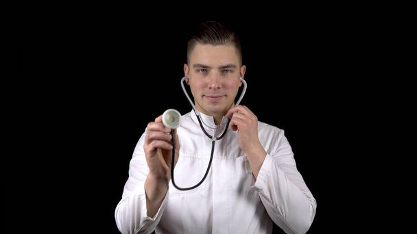A young man doctor stands with with a stethoscope and listens on a black background. The man directed the hearing aid into the camera. - Photo, Image