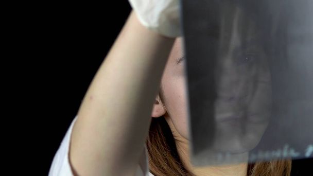 Jonge vrouwelijke arts controleert de resultaten van een röntgenfoto van de voet. Een meisje overweegt een close-up röntgenfoto. Op een zwarte achtergrond. Bekijk door een röntgenfoto. - Foto, afbeelding