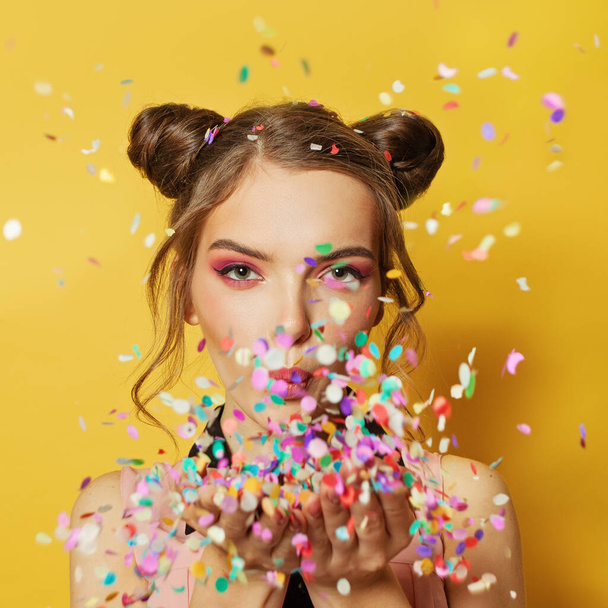Happy beautiful woman blowing colorful confetti on yellow card background - Fotografie, Obrázek