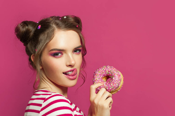 Όμορφη γυναίκα με ντόνατ και γλώσσα γλείφει τα χείλη κάποιου σε πολύχρωμο ροζ φόντο - Φωτογραφία, εικόνα