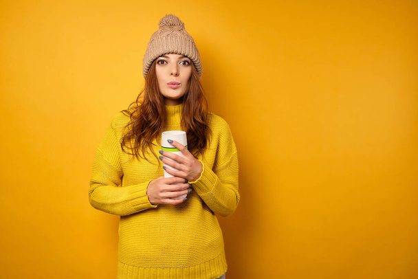 A brunette in a yellow sweater and cap with a white thermos stands on a yellow background, her lips stretched out - Foto, afbeelding