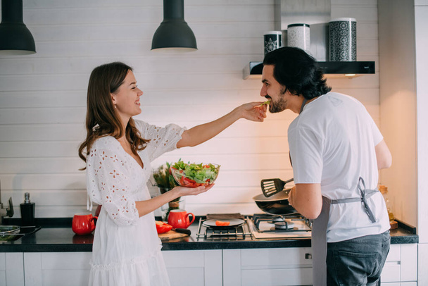 国際女性デー。自宅のキッチンで朝の若いカップル。男は美しいスタイリッシュなキッチンで3月8日の女性のお祝いの朝食を調理します. - 写真・画像