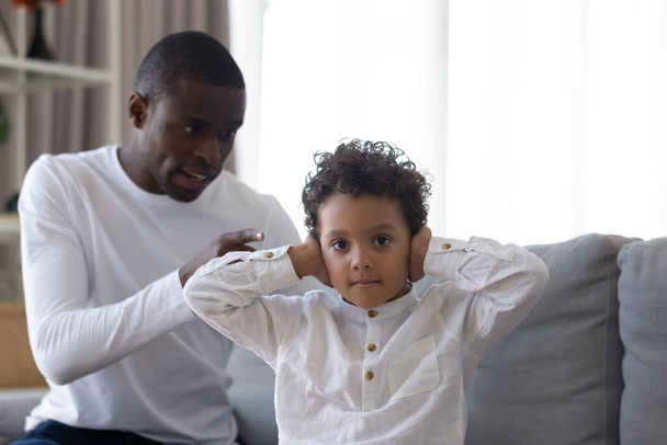 Μικρός γιος καλύπτει τα αυτιά, αγνοώντας αυστηρά θυμωμένος Αφρο-Αμερικανός πατέρας - Φωτογραφία, εικόνα