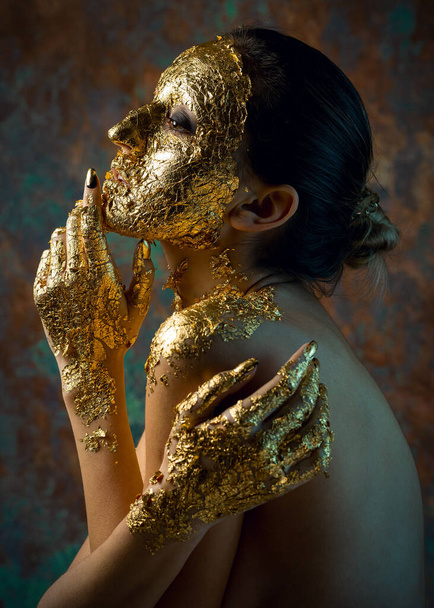 金箔で作られた顔にマスクをした女の子。グラマラスなスタジオポートレートのブルネット-抽象的な背景. - 写真・画像
