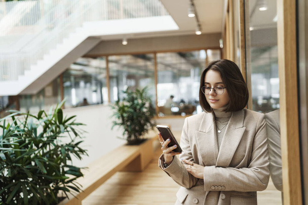 Lifestyle, women career concept. Good-looking young female entrepreneur, office lady in stylish jacket waiting for person in hall of hotel, checking messages on smartphone, smiling at phone - Zdjęcie, obraz