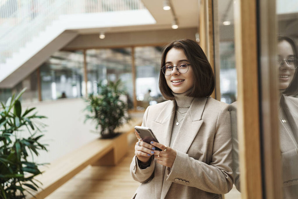 Portrait of successful happy young woman start prominent career, sending message, use mobile phone and smiling at camera as waiting for someone in office hall, reception, meeting business partners - Фото, зображення
