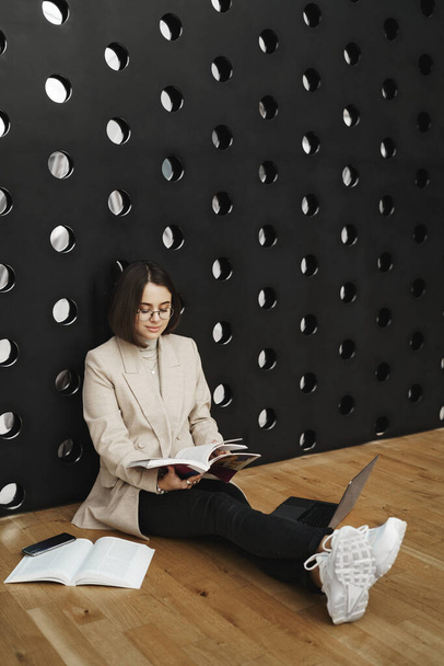 Vertical portrait of smart pretty young woman studying on floor with laptop and books, reading worksheet, learning new material, prepare report for part-time job, lean on fancy wall - Valokuva, kuva