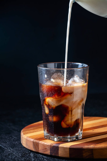 Milk Being Poured into Iced Coffee in Tall Glass on Dark Background. Concept Refreshing Summer Drink - 写真・画像