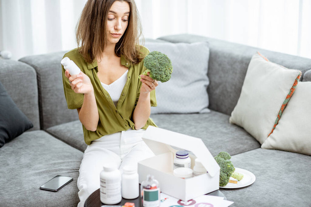 Thoughtful woman with nutritional supplements and broccoli at home - Photo, image