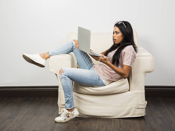 Young woman with her legs up on a couch, with a surprise by what is happening on a laptop screen - Photo, Image