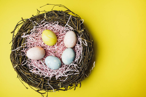 Egy gallyakból és szénából álló fészek és egy rózsaszín papírtöltelék pasztell színű húsvéti tojásokkal. Színes tojások a fény ünnepe húsvét. Húsvéti kártya sárga alapon. - Fotó, kép
