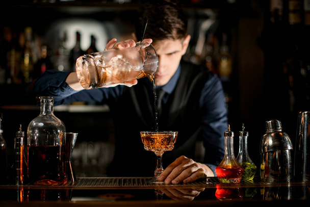 Young bartender gently pours brown cocktail into glass. - Фото, зображення