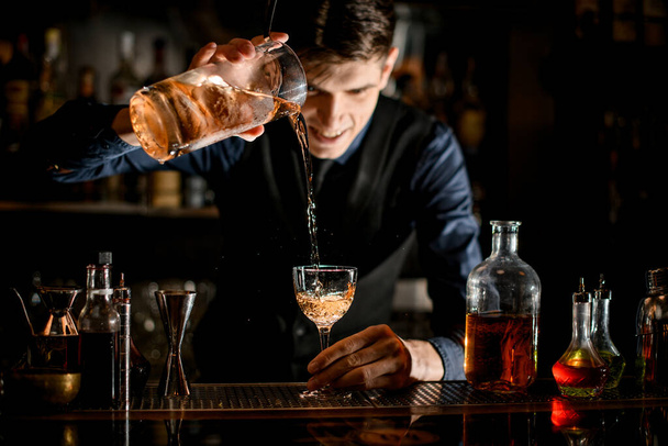 Bartender holds in one hand glass with a cocktail and pours it into wineglass in his other hand. - Φωτογραφία, εικόνα