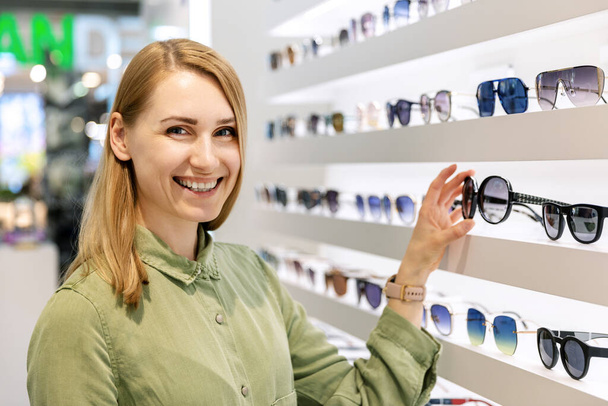 mujer sonriente eligiendo gafas de sol de estante en la tienda de gafas
 - Foto, Imagen