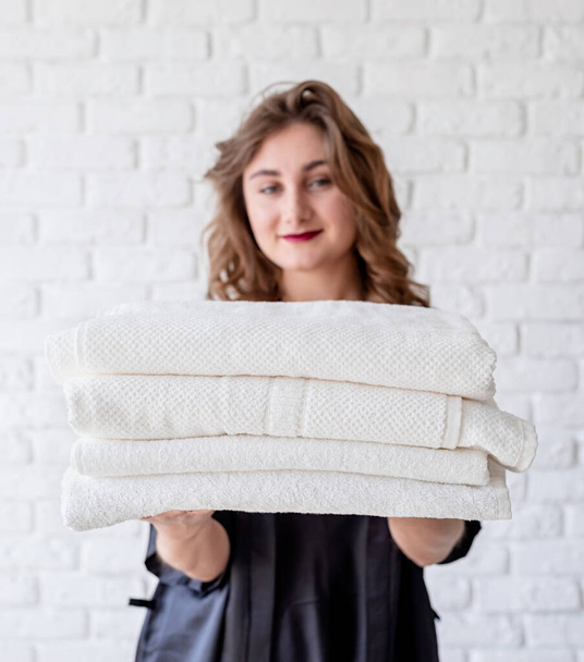 sonriente joven mujer sosteniendo una pila de toallas sobre fondo de ladrillos blancos
 - Foto, imagen