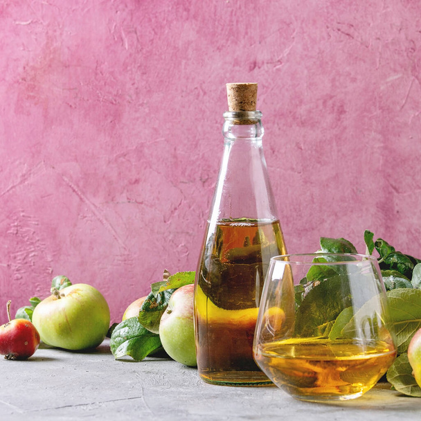 Bottle ang glass of apple juice with garden apples with leaves and branches on table with pink background. Autumn home harvesting. - Фото, зображення