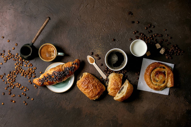 Variety of traditional french puff pastry raisin and chocolate buns, croissant with various cups of coffee and milk, cezve, recycled wooden spoon of sugar over dark texture background. Flat lay, space - Foto, immagini