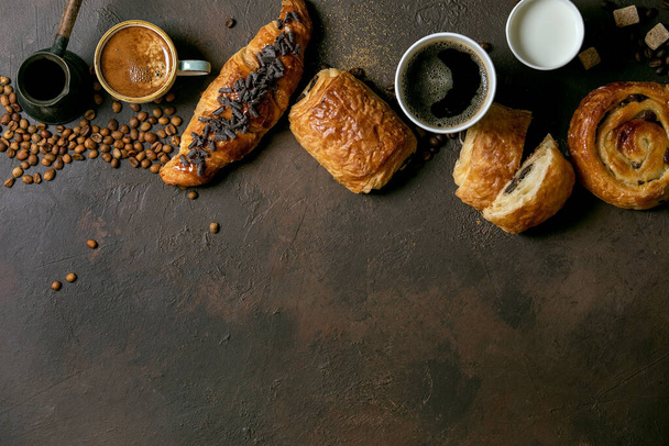 Variety of traditional french puff pastry raisin and chocolate buns, croissant with various cups of coffee and milk, cezve, recycled wooden spoon of sugar over dark texture background. Flat lay, space - Foto, Imagen