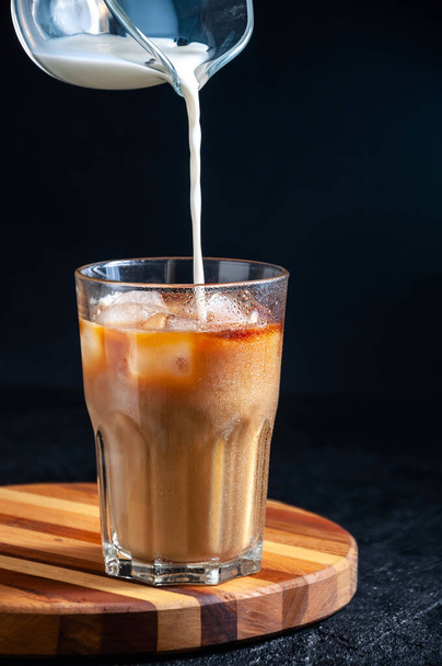 Молоко наливают в ледяную кофейню в высоком стекле на темном фоне. Концепция освежающего летнего напитка - Фото, изображение