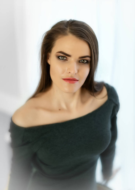 Close-up portrait of young pretty female 20-30 years old dressed in a feminine sexy dress with an open neckline standing next to window with white curtains - Fotoğraf, Görsel