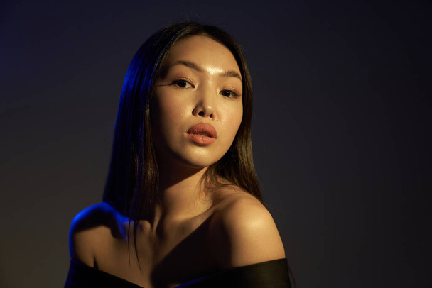 Portrait d'une jeune belle asiatique regardant directement la caméra. Les cheveux et les épaules de la femme dans des lumières bleu UV lumineux colorés. Sur fond noir
.                                   - Photo, image