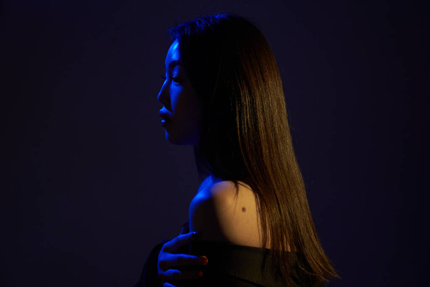  Profile of a young beautiful Asian girl, looking away. A woman's face in colorful bright UV-blue lights. On a gray background.                                  - Foto, imagen