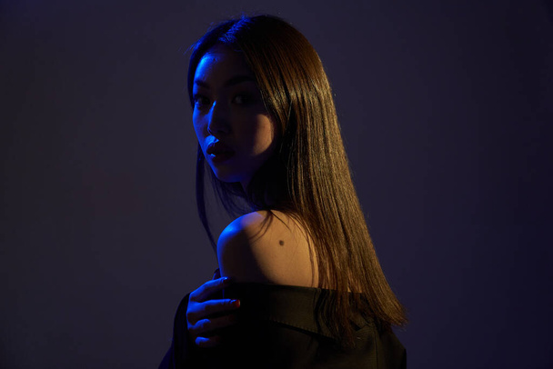  Portrait of a young beautiful Asian girl looking at the camera. A woman's face and shoulders in colorful bright UV-blue lights. On black backgrounds.                                - Foto, imagen
