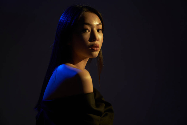   Portrait of a young beautiful Asian girl looking directly at the camera. A woman's back in colorful bright UV blue lights. On black background.                                  - Foto, afbeelding
