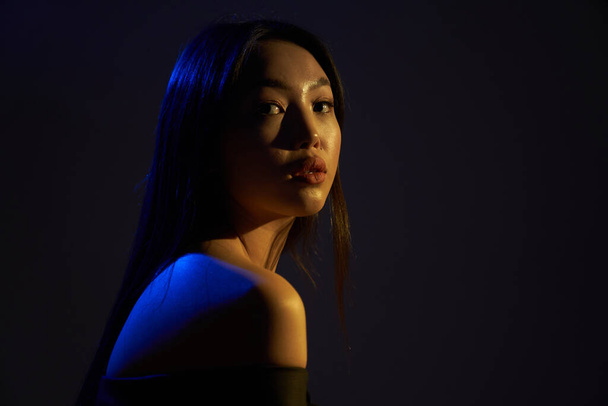 Profile of a young beautiful Asian girl, she looks directly at the camera. The hair and the back of the woman in bright , bright blue ultraviolet lights. On black background.                                - Foto, afbeelding