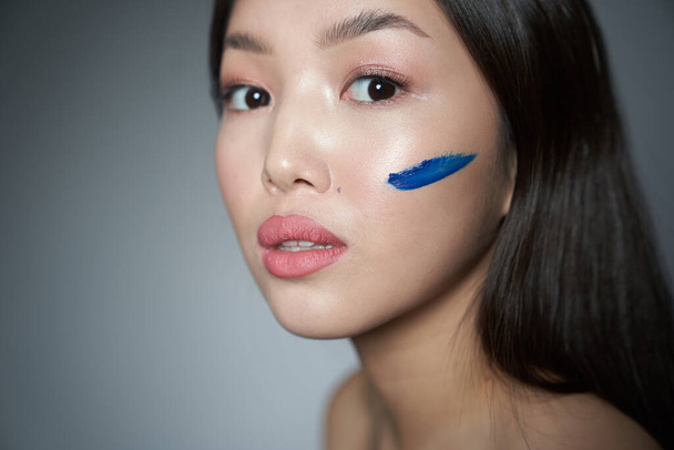 Portrait of a young beautiful Asian girl with smeared oil blue paint, cheek in the form of a horizontal strip. Looks directly at the camera. On a gray background, a place to copy. The concept of creat                                - Photo, Image
