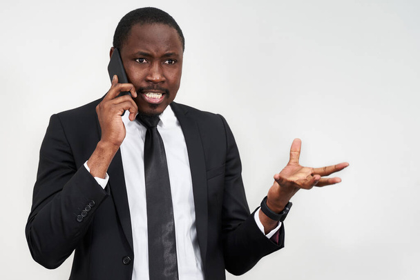 Nahaufnahme eines wütenden jungen Afrikaners, der schreit, während er auf dem Smartphone vor grauem Hintergrund spricht - Foto, Bild