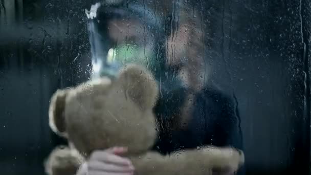 Bezorgd kind draagt gasmasker terwijl hij Teddy Bear strak vasthoudt. Het is donker en de regen stroomt uit het raam..  - Video