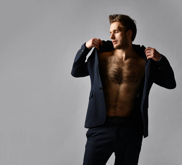 Handsome man in black classic suit on naked body. He is straightening his jacket, posing against gray background. Close up - Foto, afbeelding