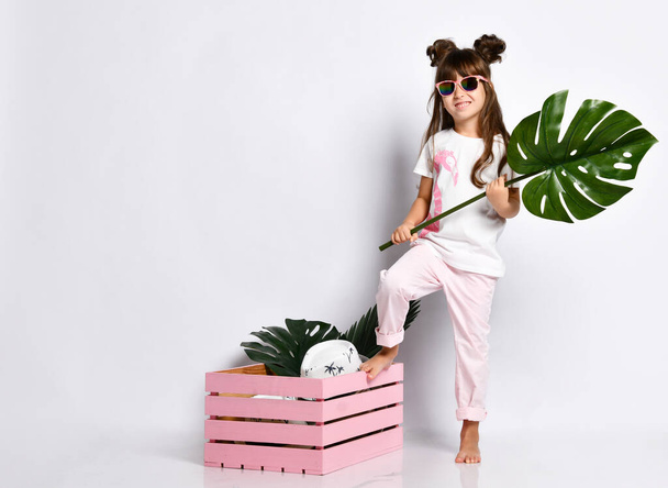 A beautiful girl poses standing next to a pink wooden box, wearing A Flamingo t-shirt and sunglasses with a leaf from a flower in her hands, barefoot - Photo, image