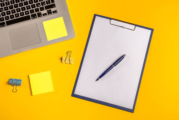 フラットレイアウト。紙クリップ、フォリオ、ペンと付箋で黄色の背景にコンピュータ。ビジネスコンセプトのアイデア。スペースのコピー. - 写真・画像