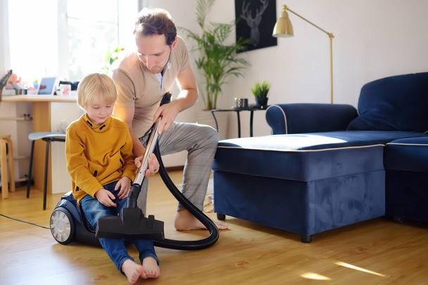 Young handsome father teaches his son how to use a vacuum cleaner. Loving dad and his preschooler boy doing housework together at home. Happy fatherhood - 写真・画像