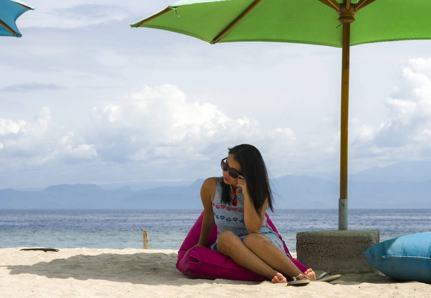 joven hermosa y feliz mujer china asiática en elegante vestido de verano y gafas de sol posando relajado en la playa tropical relajado sentado en bolsa de frijoles bajo sombrilla
 - Foto, Imagen