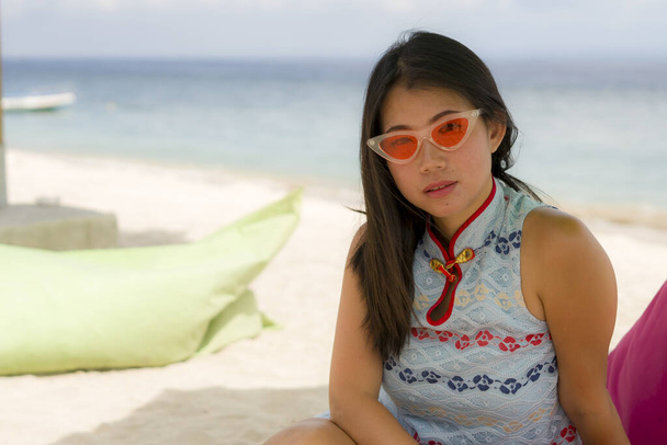 young beautiful and happy Asian Korean woman in chic Summer dress and hipster sunglasses posing relaxed at tropical beach relaxed sitting on beanbag under umbrella parasol - Zdjęcie, obraz