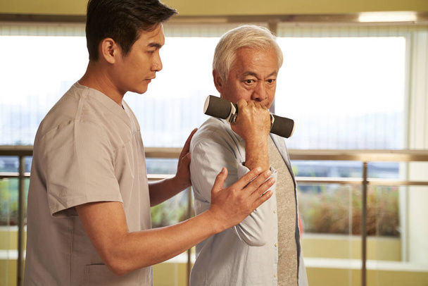senior asiático hombre ejercitando usando dumbbells guiado por físico terapeuta en rehabilitación centro
 - Foto, Imagen